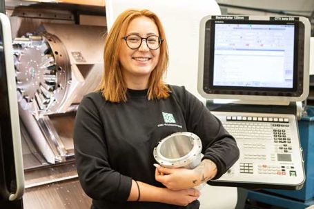 Mitarbeiterin mit Werkstück vor der CNC-Drehmaschine
